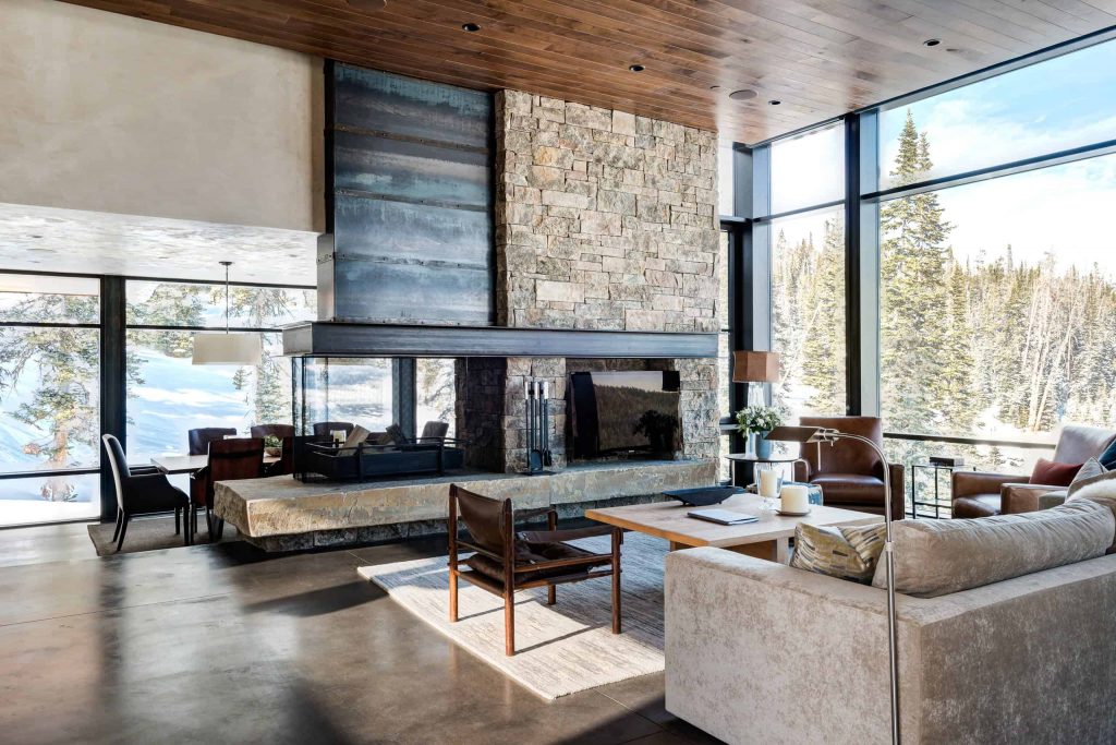 Spacious living room in a house with big tile floor, sofa and chairs, a fireplace in a stone wall in the middle of the room and a TV next to the fireplace