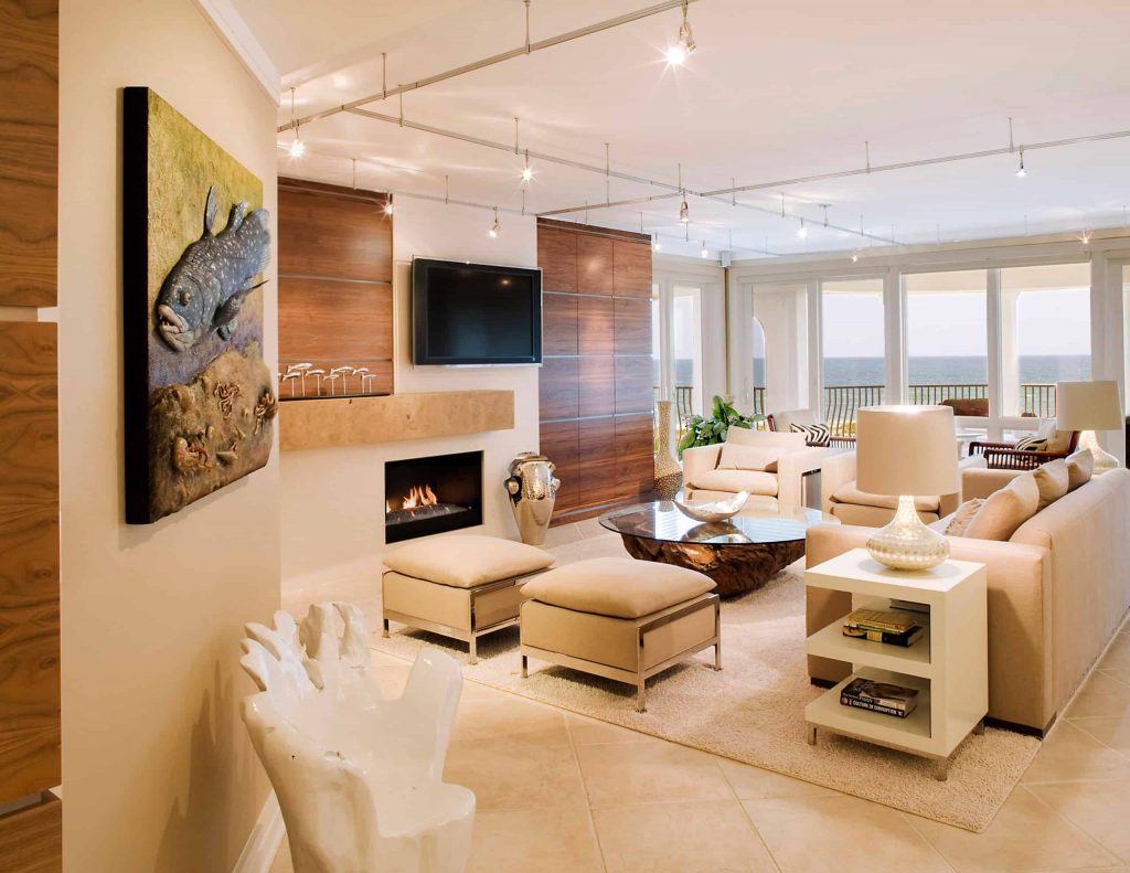 Spacious living room in beige colors in a house with tile floor, wooden panels on the walls, sofa, chairs and ottomans, a balcony with a view of the sea, a small fireplace and a TV above the fireplace