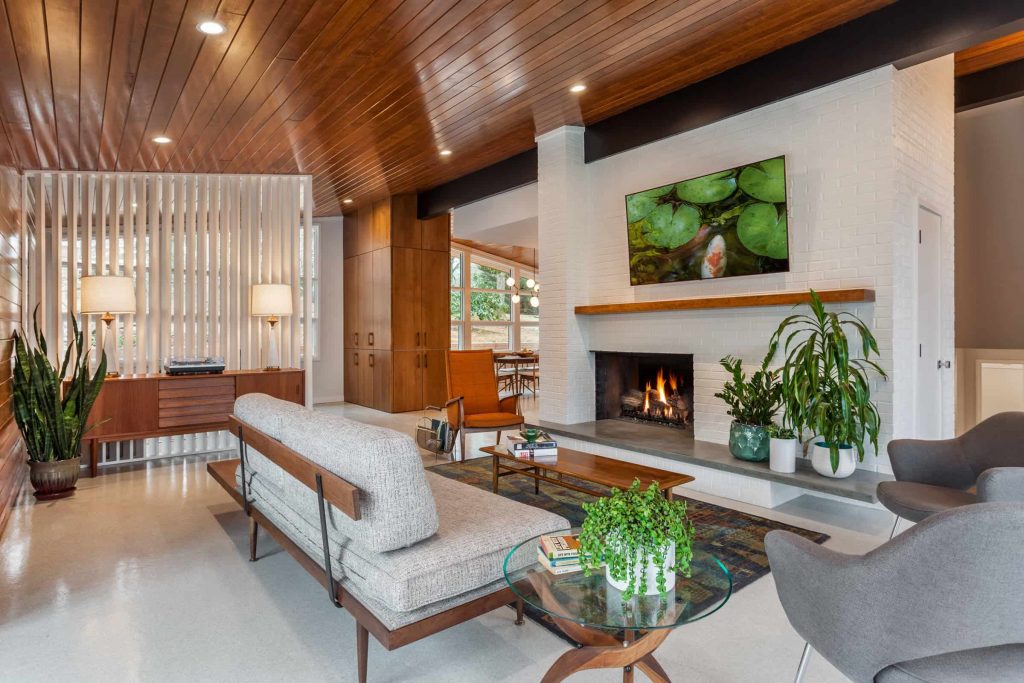 Spacious living room in a house with wooden ceiling and cabinets, grey sofa and chairs, a white brick fireplace and a TV above the fireplace