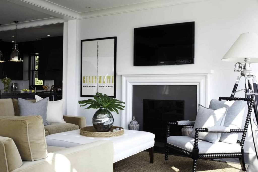 Small living room next to the kitchen in a house with white walls, brown sofa and grey chair, grey fireplace, big poster next to the fireplace and a TV above the fireplace