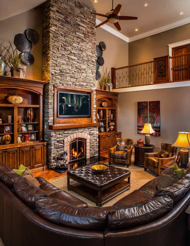 Rustic living room with brown leather sofa, chairs, cupboards, stone fireplace with a chimney and a TV in a wooden frame above the fireplace