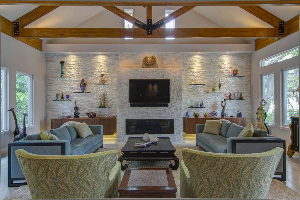 Spacious contemporary living room with white walls, blue sofas and colurful armchairs, white stone tile wall with vases on shelves, wide black fireplace and a TV above the fireplace
