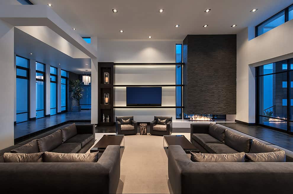 Spacious contemporary living room with white walls, dark brown sofas and armchairs, wide fireplace under a dark brown stone tile chimney and a TV next to the fireplace