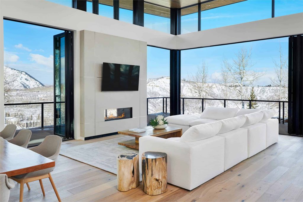 Spacious contemporary living room with a balcony and window walls, wooden floor, white walls and sofa, small fireplace and a TV above the fireplace