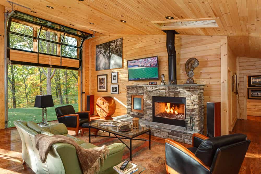Small rustic living room with wooden floor, walls and ceiling, window wall, green sofa, black armchairs, small stone tile fireplace with a chimney and a TV above the fireplace